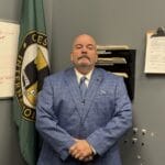 Picture of a man (Director Ray Strobel) wearing a suit standing in front of a Chesley Brown Flag.