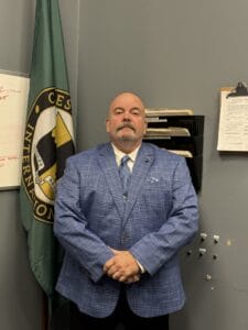 Picture of a man (Director Ray Strobel) wearing a suit standing in front of a Chesley Brown Flag.