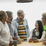 A vibrant, multigenerational team shares laughter and conversation over coffee in a naturally lit, relaxed office setting. Workplace safety culture, safety leadership, employee engagement in safety, accountability in workplace safety